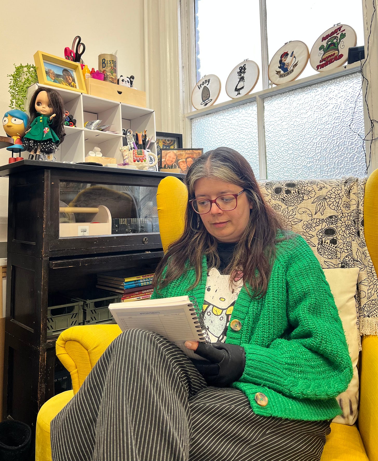 Annie wearing a green cardigan sits writing in her yellow armchair at her art studio surrounded by art supplies, books & art work