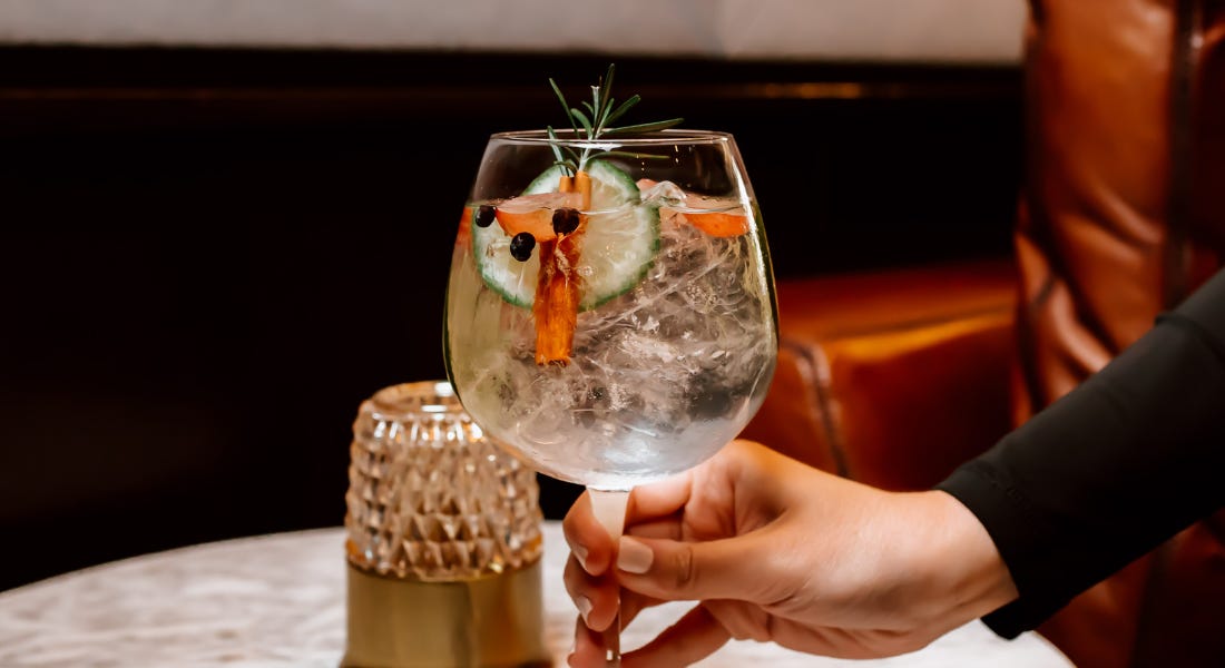 A hand holding a large gin glass with cucumber and rosemary in it, pictured on top of a marble table