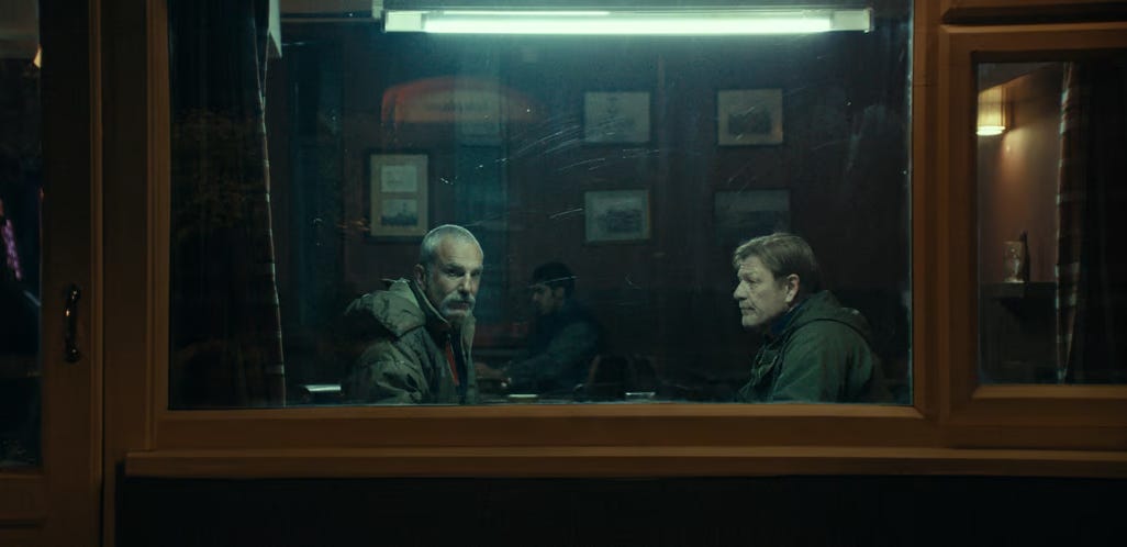 Two men sit across from one another, by a window, in a bar. The light above them is a hazy blue. One gazes out of the window, towards the camera and viewer. It is dark.