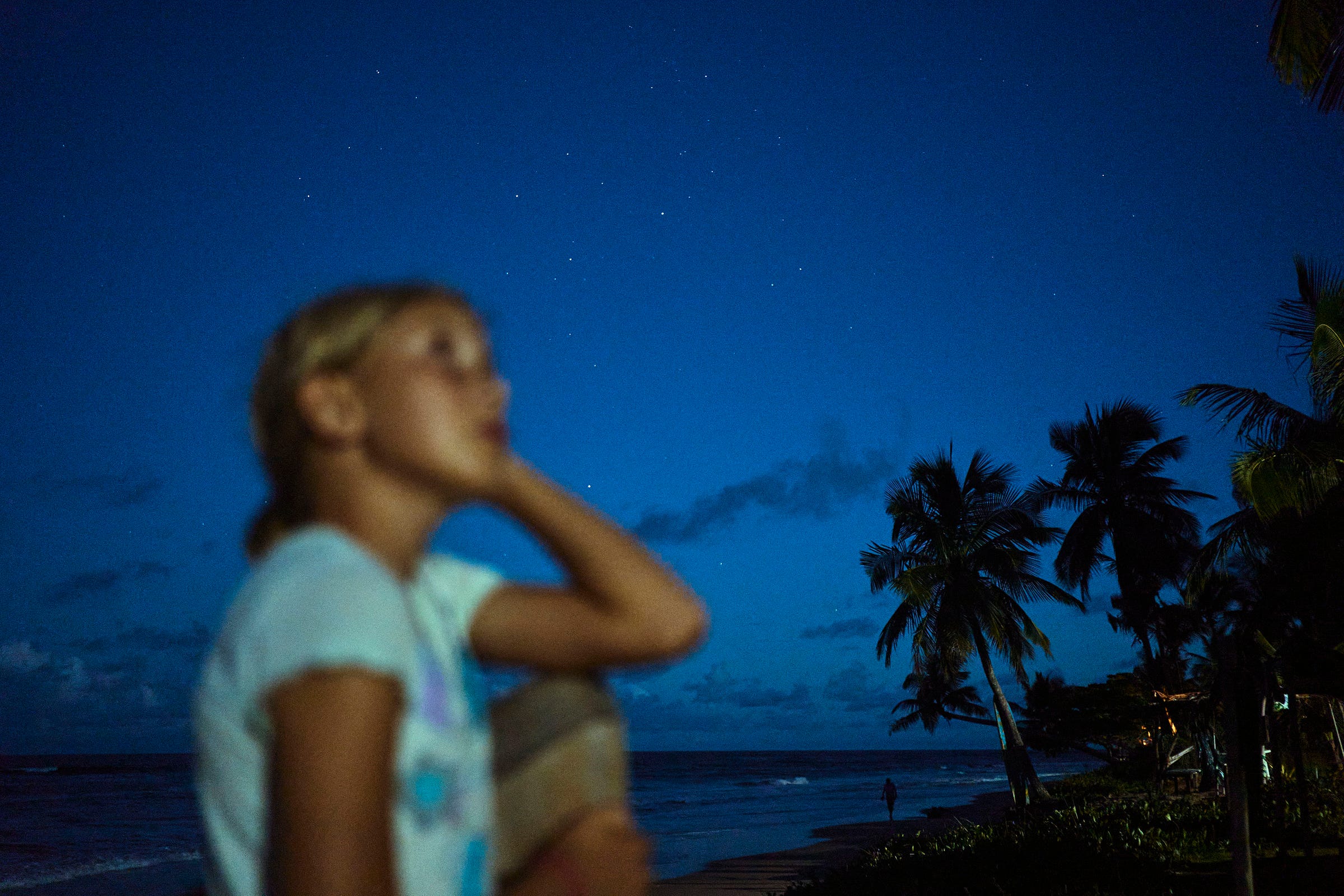 A blonde girl sits on a beach at night, looking upward. Palm trees and the ocean are in the background under a dark, starry sky with scattered clouds. The child is blurred in the foreground.
