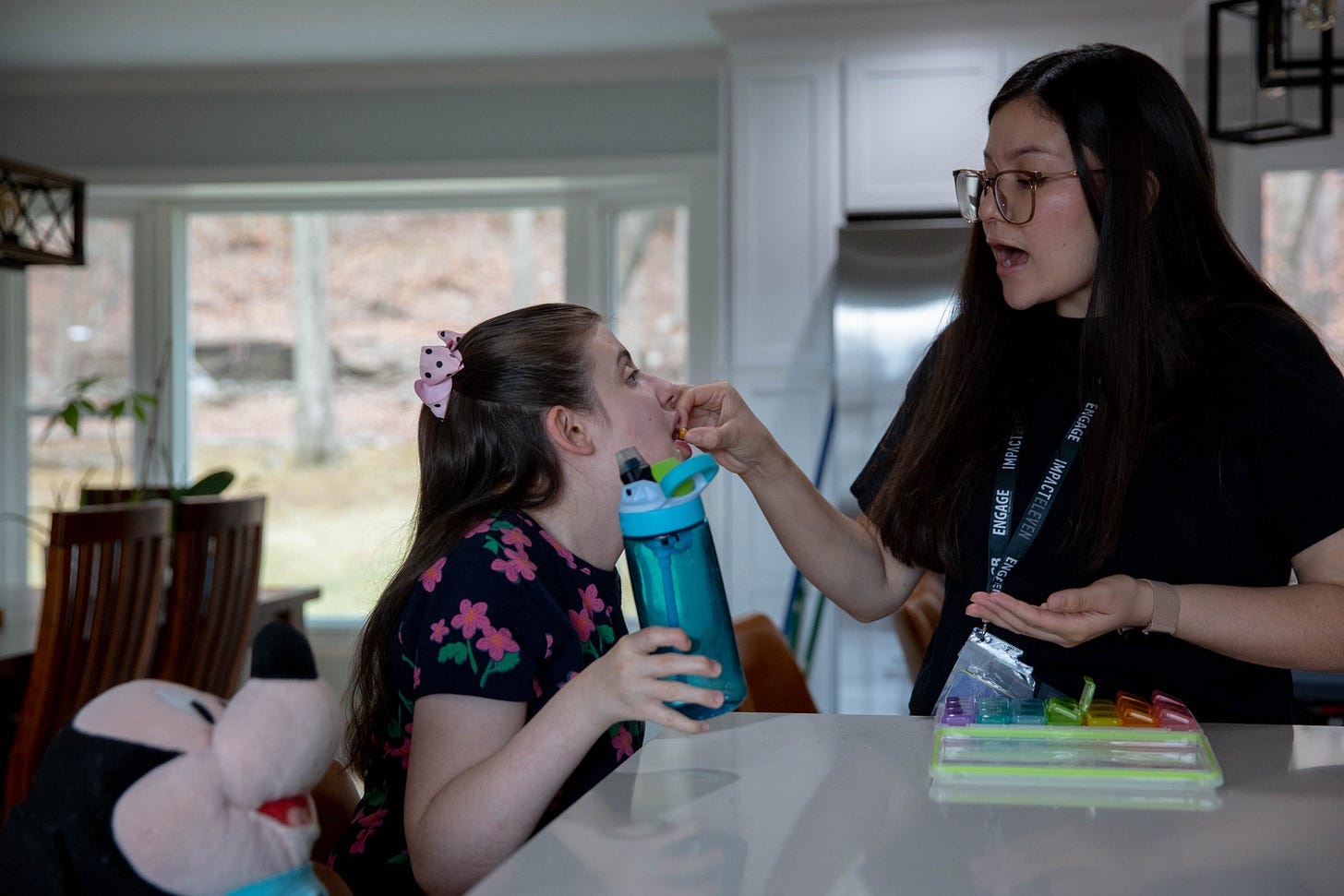 A woman with long brown hair wearing glasses puts a pill into the mouth of a girl sitting at a countertop with her mouth open A woman with long brown hair wearing glasses puts a pill into the mouth of a girl sitting at a countertop with her mouth open