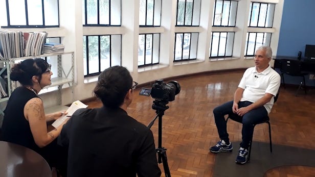 #descrevemospravocê  A imagem mostra uma entrevista sendo gravada em um ambiente interno, com piso de madeira e muitas janelas retangulares ao fundo, que permitem a entrada de luz natural. À direita, um homem de cabelos grisalhos, pele clara e expressão tranquila está sentado em uma cadeira preta, vestindo camiseta polo branca, calça escura e tênis azul. Ele olha em direção às pessoas que o entrevistam.  À esquerda da imagem, uma mulher e um homem estão voltados para o entrevistado. A mulher tem cabelos escuros presos em um coque, usa brincos e uma blusa preta sem mangas, com tatuagens visíveis no braço esquerdo. Ela segura um caderno ou bloco de notas e sorri enquanto conversa. O homem está de costas para a câmera e usa roupa escura, com o cabelo preso. Entre eles, há uma câmera profissional montada sobre um tripé, posicionada para gravar o entrevistado.  À esquerda, ao fundo, há uma estante metálica com vários livros e documentos organizados. Do lado direito, próximos à parede azul, é possível ver computadores sobre mesas. O ambiente lembra uma biblioteca ou espaço institucional de pesquisa e registro de memória.