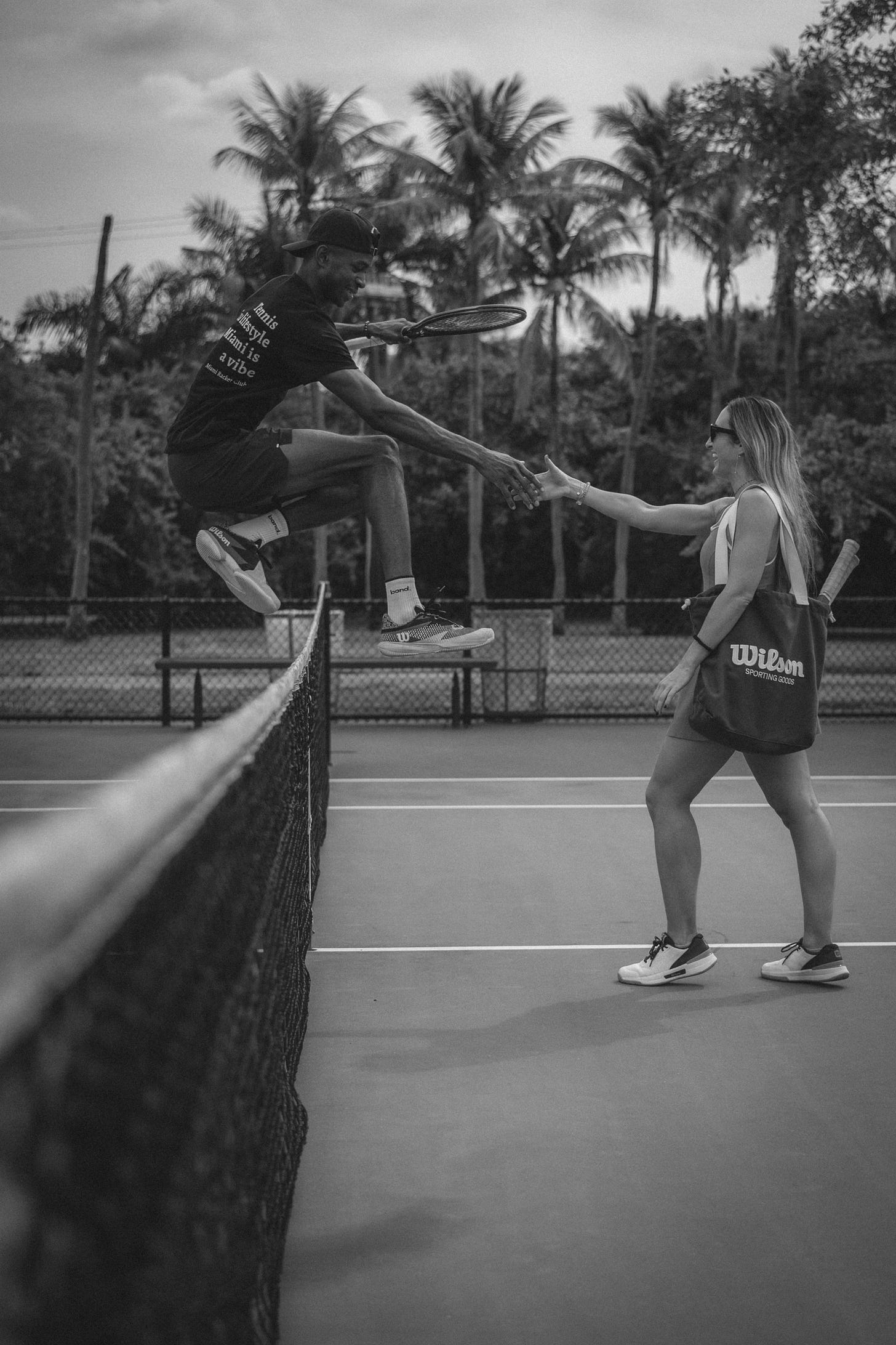 Black and white photo of Sibi Soumahoro mid-jump over a tennis net, reaching to shake hands with Clara Pérez, who stands smiling in surprise. Clara holds a Wilson tote with a racquet; palm trees and court fencing in the background. Captures a spontaneous, lighthearted post-match moment. Black and white photo of Sibi Soumahoro mid-jump over a tennis net, reaching to shake hands with Clara Pérez, who stands smiling in surprise. Clara holds a Wilson tote with a racquet; palm trees and court fencing in the background. Captures a spontaneous, lighthearted post-match moment.