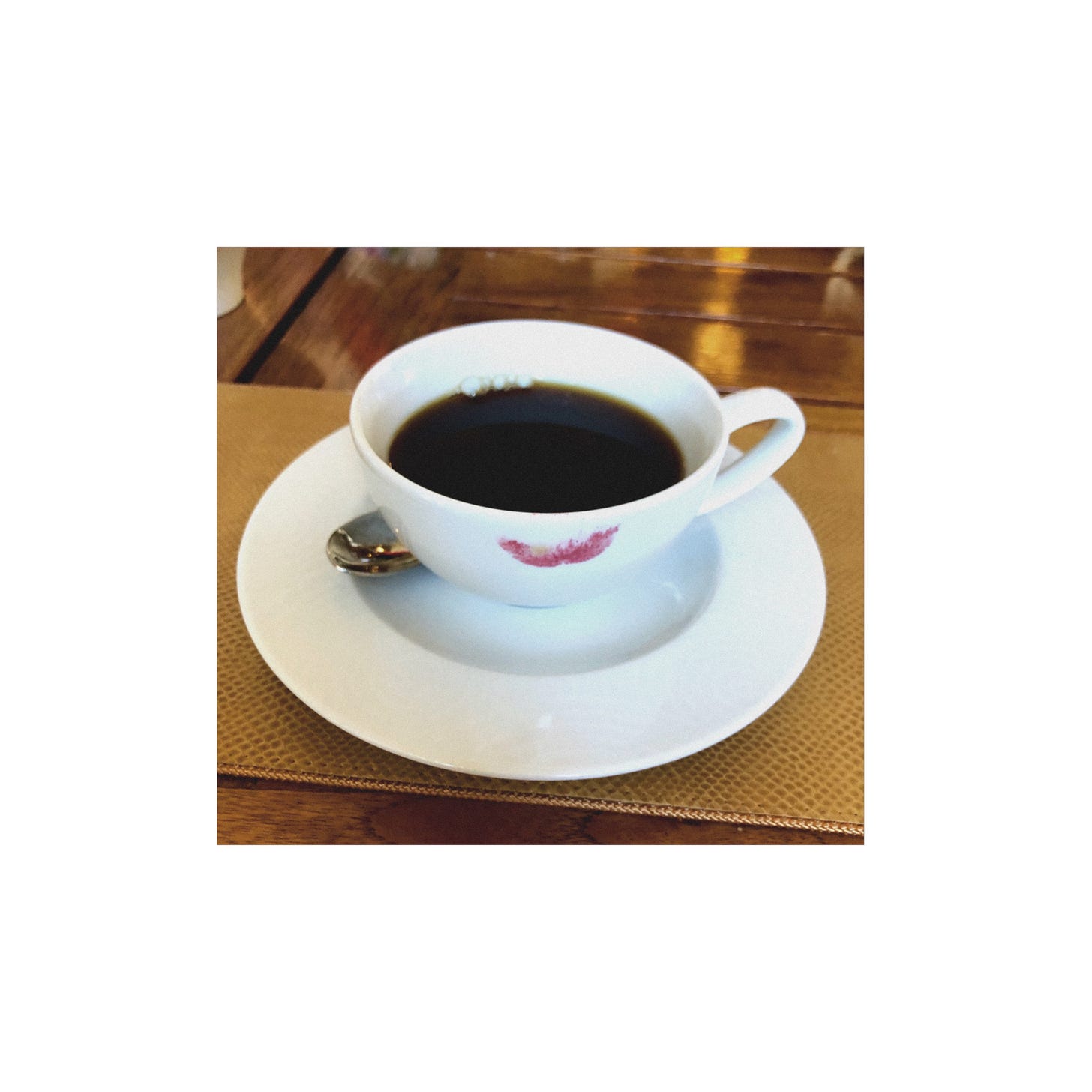 black coffee in a white porcelain cup on a white porcelain saucer, a pinkish lipstick mark on the cup, with a spoon tucked behind the cup 