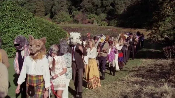 Yellow-tinged film still of a long procession of people wearing ragged animal masks that have seen better days. They are wearing kilts, long 70s dresses (it was the 70s). They walk along a groomed grass path with a woodland behind them.