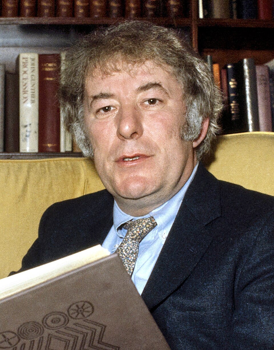 Head and shoulder shot of Heaney in 1982; he’s a white man with unkempt graying hair and a big face; wearing a coat and patterned tie; he is on a couch reading a book; behind him is a full bookcase 