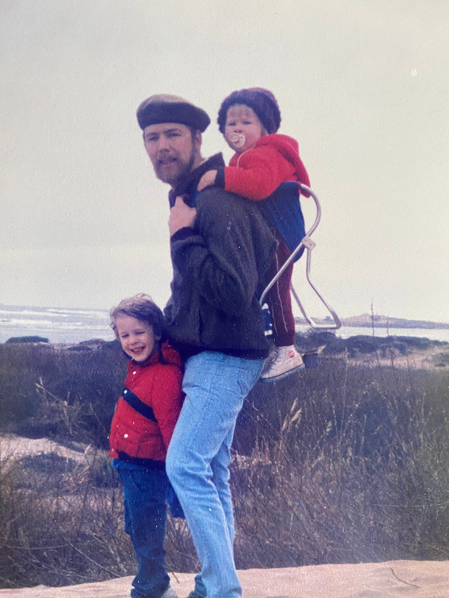 A young Cormac rides in a framed backpack carrier on his dad's back while his brother leans affectionately against their dad's leg during a coastal outing.