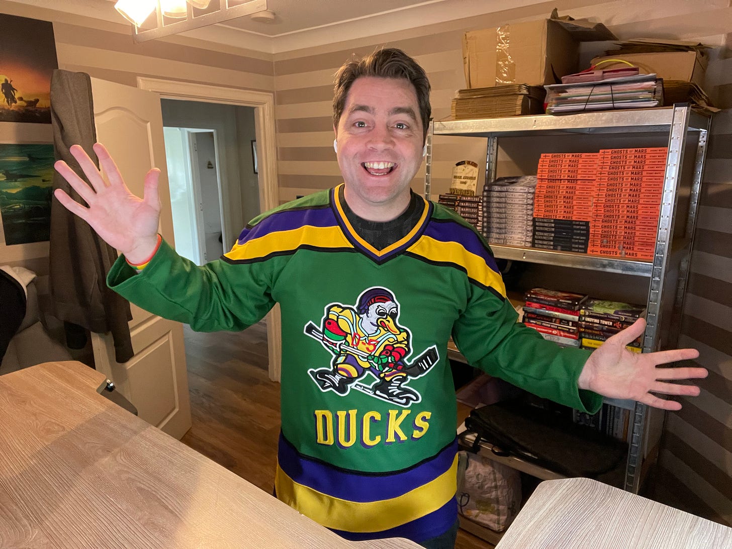 Stuart smiling at camera with arms wide, wearing a Might Ducks replica hockey shirt.