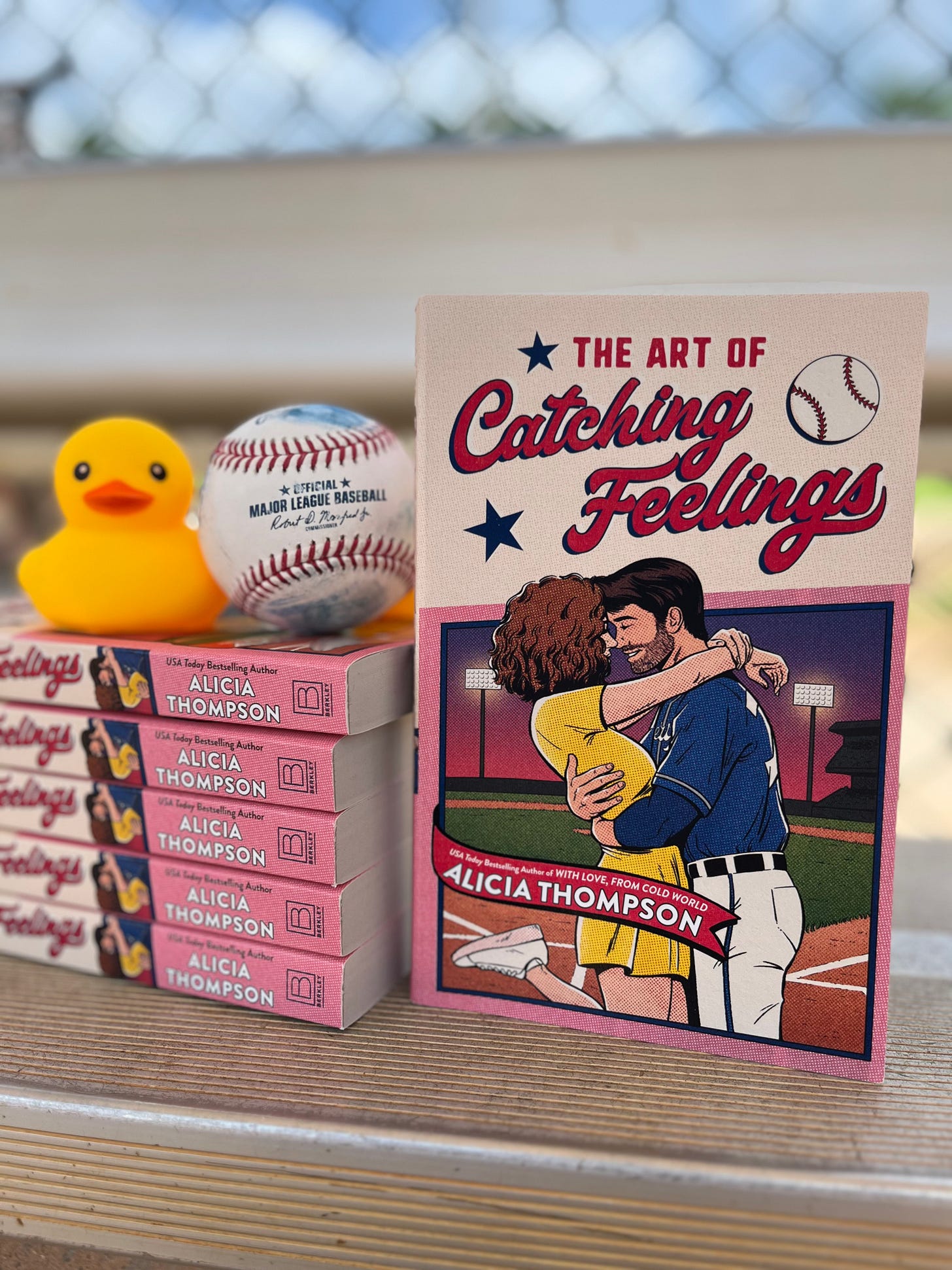 Photo of a stack of THE ART OF CATCHING FEELINGS with a yellow duckie and a baseball resting on top of them, and then a copy propped up next to the stack with the cover out, all taken on a bench in a Little League dugout with the fence behind