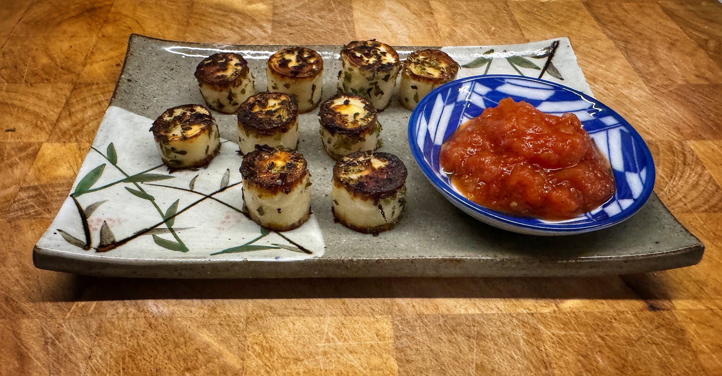 9 seared hearts of palm "scallops" presented on a rectangular Japanese serving dish. There's also a small ceramic bowl holding chili sauce on the serving dish. 9 seared hearts of palm "scallops" presented on a rectangular Japanese serving dish. There's also a small ceramic bowl holding chili sauce on the serving dish.