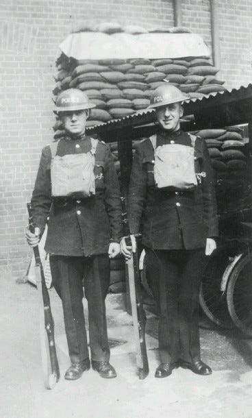 Two officers stand in uniform, complete with satchels and tin hats.