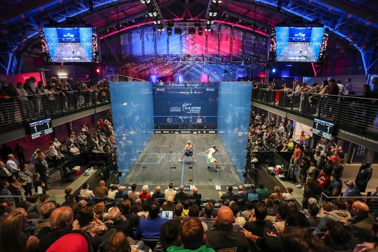 Two women athletes compete on squash court with four transparent walls surrounded by onlookers