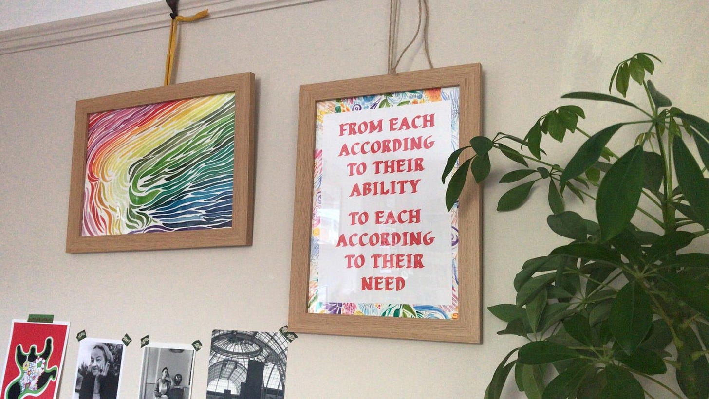 A poster of the words 'from each according to their ability to each according to their need' with a colourful border, then another rainbow poster, an umbrella plant, and postcards of niki de st phalle's work, barbara hepworth, frida khalo