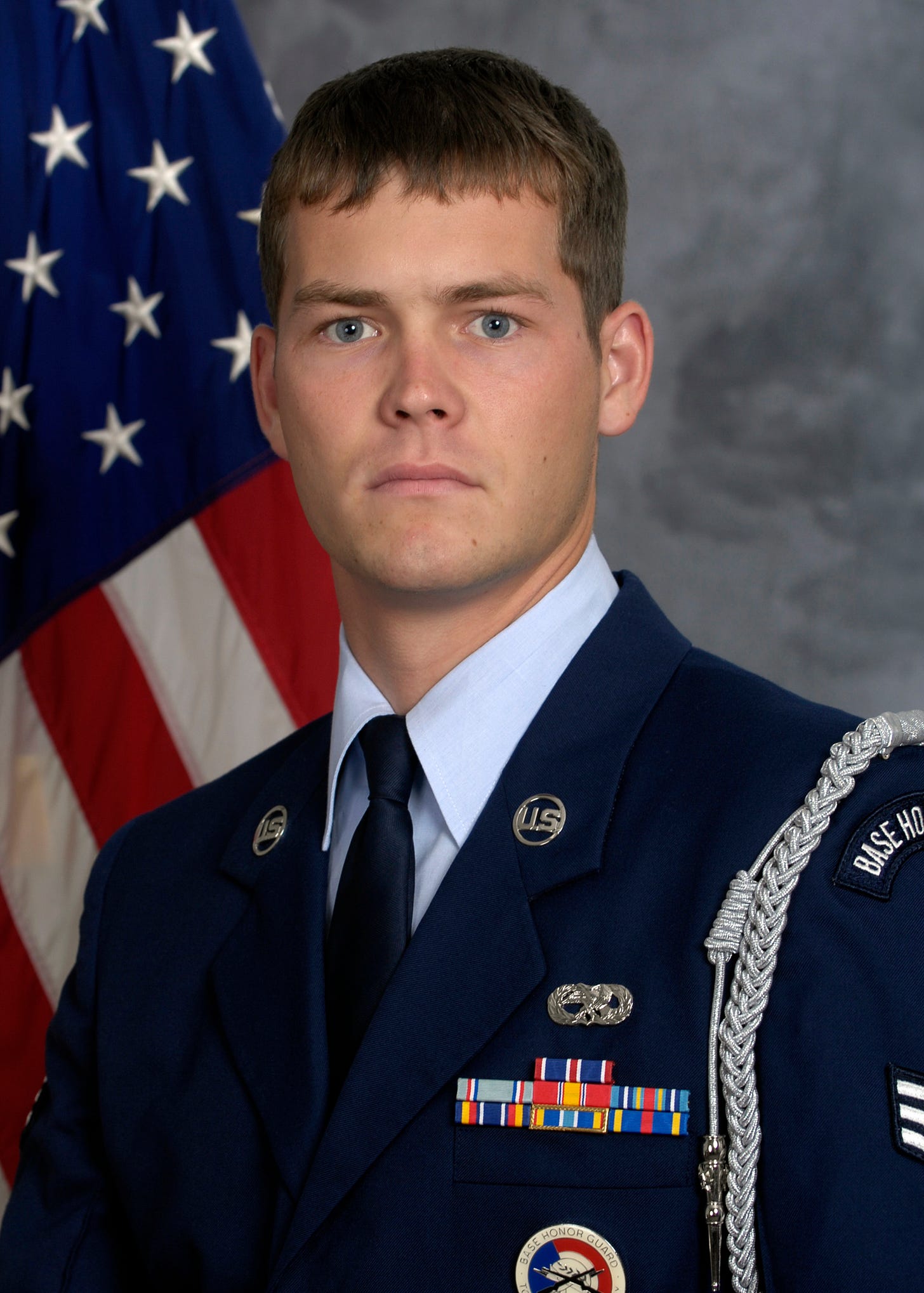 A formal portrait of a young U.S. Air Force service member in a dark blue dress uniform with ribbons, badges, and a silver shoulder cord, posed in front of a partially visible American flag and a gray studio backdrop. A formal portrait of a young U.S. Air Force service member in a dark blue dress uniform with ribbons, badges, and a silver shoulder cord, posed in front of a partially visible American flag and a gray studio backdrop.
