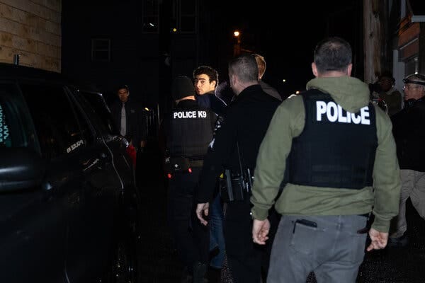 Luigi Mangione, identified by the authorities as a “person of interest” in the fatal shooting in Manhattan of a health insurance executive, looks back toward the camera as he is escorted by officers wearing vests marked “Police” and “Police K9.”