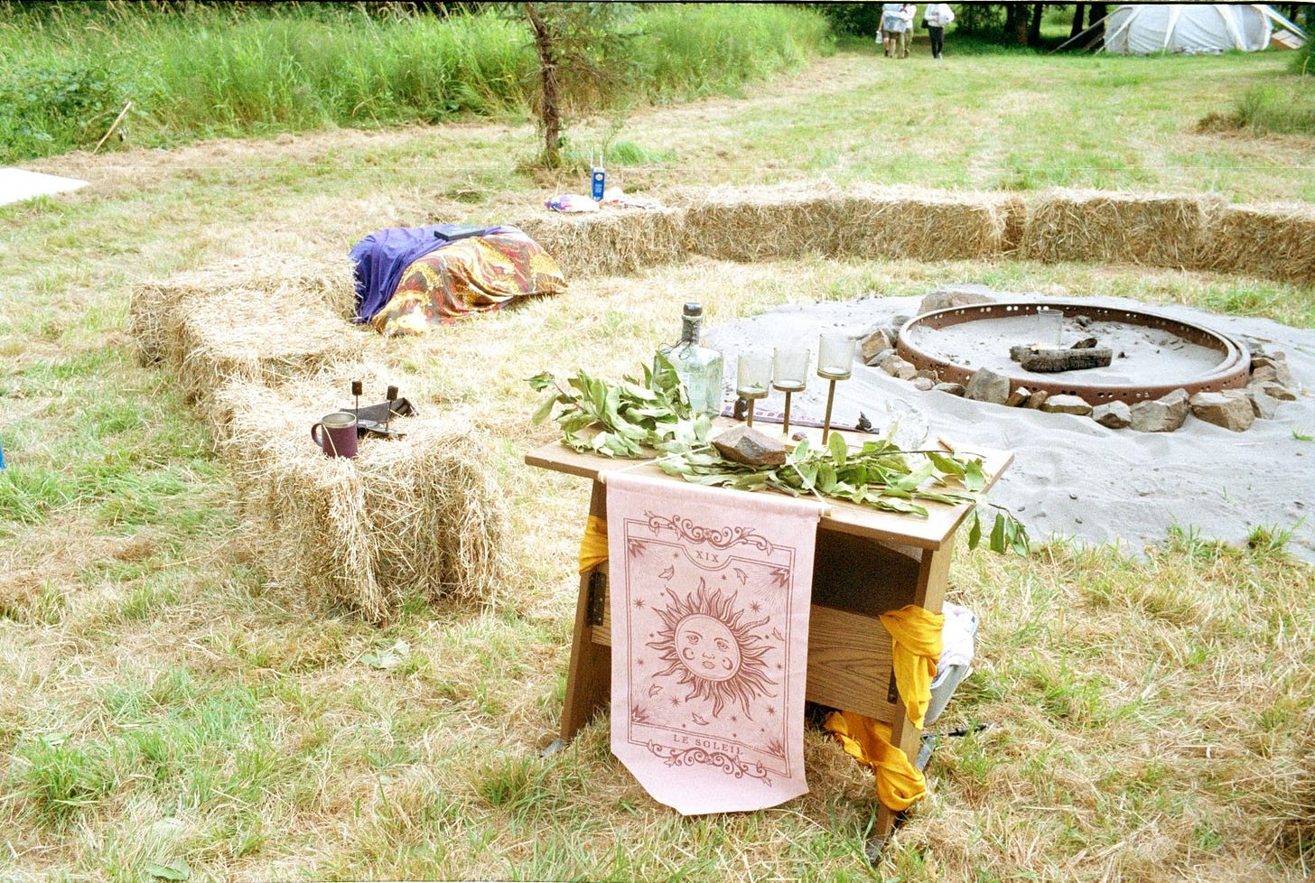 An altar to Apollo in front of some hay bales arranged in a large circle around a campfire pit. The altar has yellow cloths and a Sun tapestry and bay laurel and other Apollonian offerings. An altar to Apollo in front of some hay bales arranged in a large circle around a campfire pit. The altar has yellow cloths and a Sun tapestry and bay laurel and other Apollonian offerings.