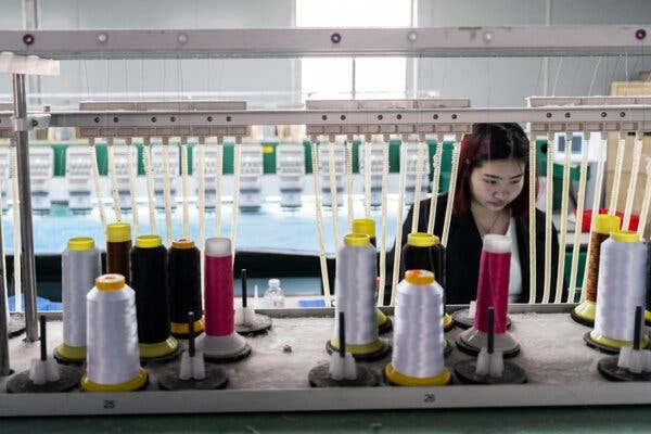 A worker stands behind rows of spools of colored thread in a factory.