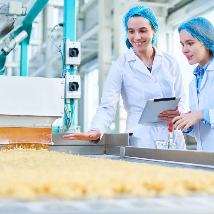 Two women in white coats and blue hairnets checking food.