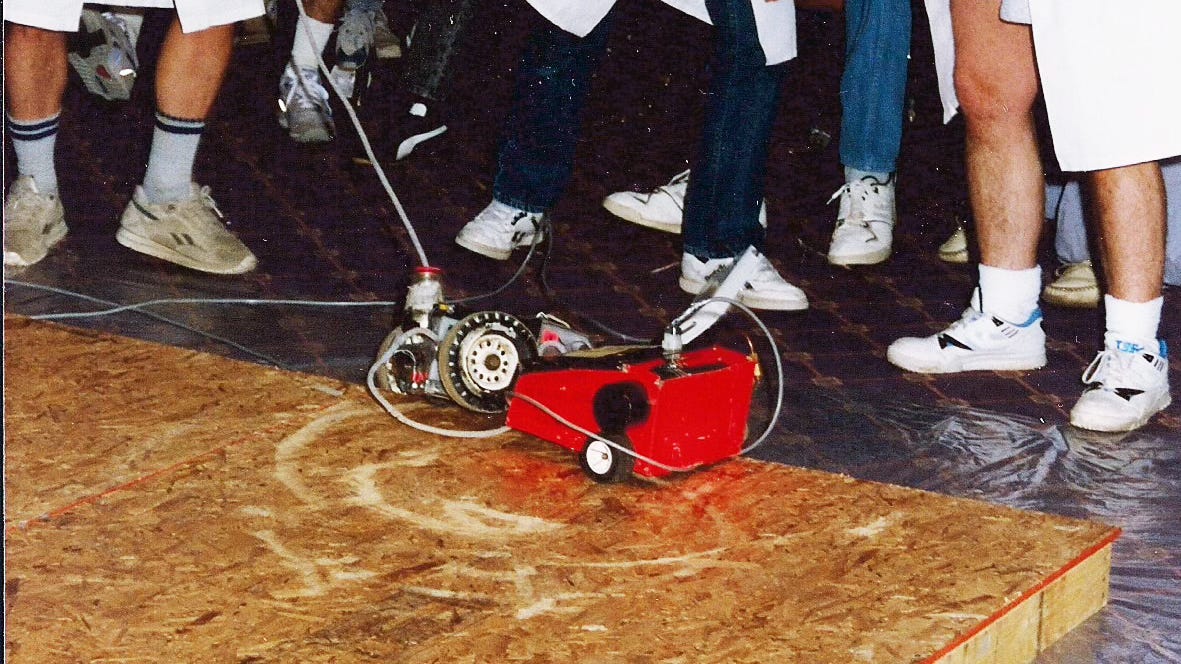 A tight crop of a grainy old photo of members of the Denver Mad Scientist Club engaging in remote-controlled robot combat at the 1989 MileHiCon. Participants wear lab coats, and two robots battle it out on a sheet of wood on the floor. A tight crop of a grainy old photo of members of the Denver Mad Scientist Club engaging in remote-controlled robot combat at the 1989 MileHiCon. Participants wear lab coats, and two robots battle it out on a sheet of wood on the floor.