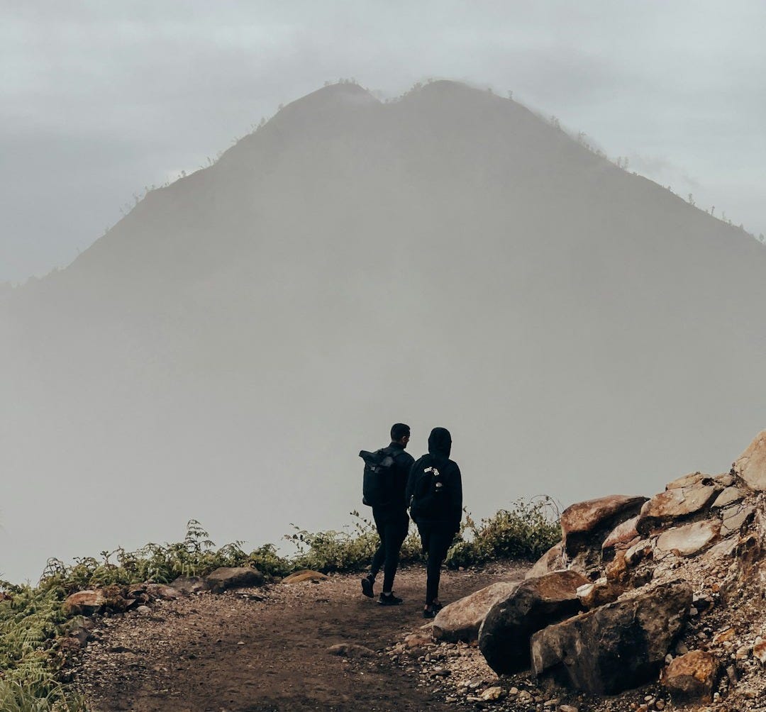 two people standing on a dirt path with a mountain in the background