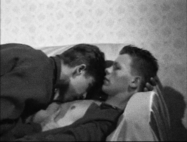 This may contain: black and white photograph of two men laying on a bed kissing each other with their noses close together