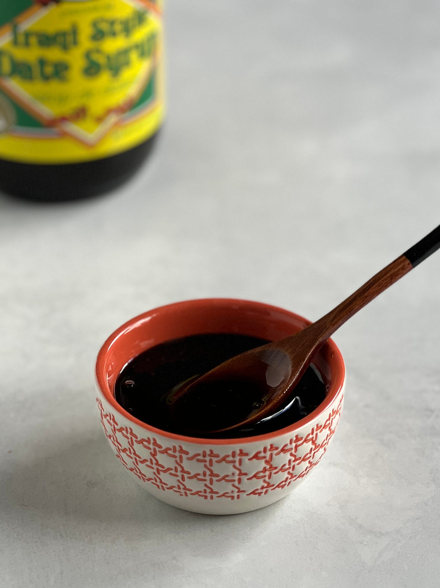 A small orange bowl of date syrup with a wooden spoon in it and a jar of date syrup in the background.