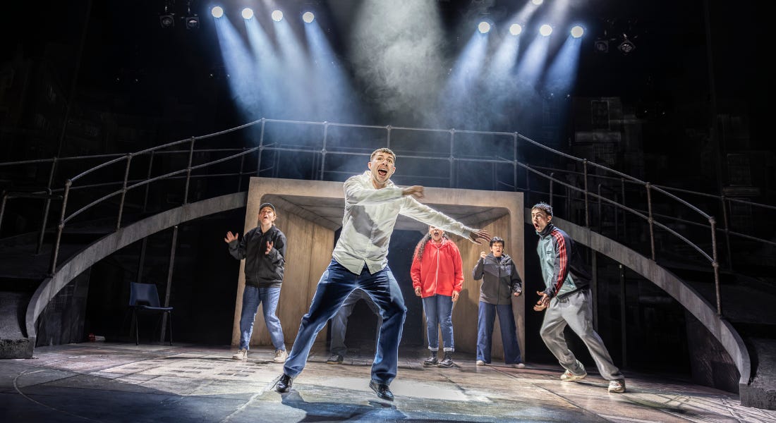 A stage set with sweeping ramps down both sides and strong spotlights. 6 actors fill the stage and are caught mid-action. The man at the front seems to be shouting.