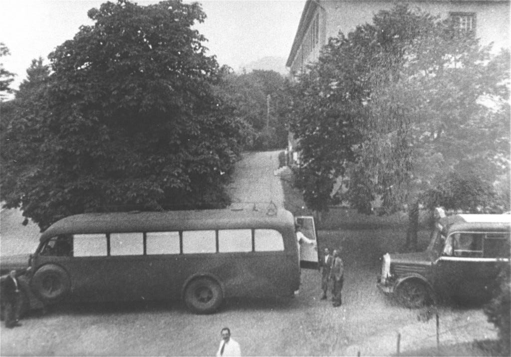 Two transport buses with painted windows are parked out front of a building with large trees. Two people stand at the back of the first bus and converse with a person opening the door at the back of the bus. All individuals appear to have light skin tones.