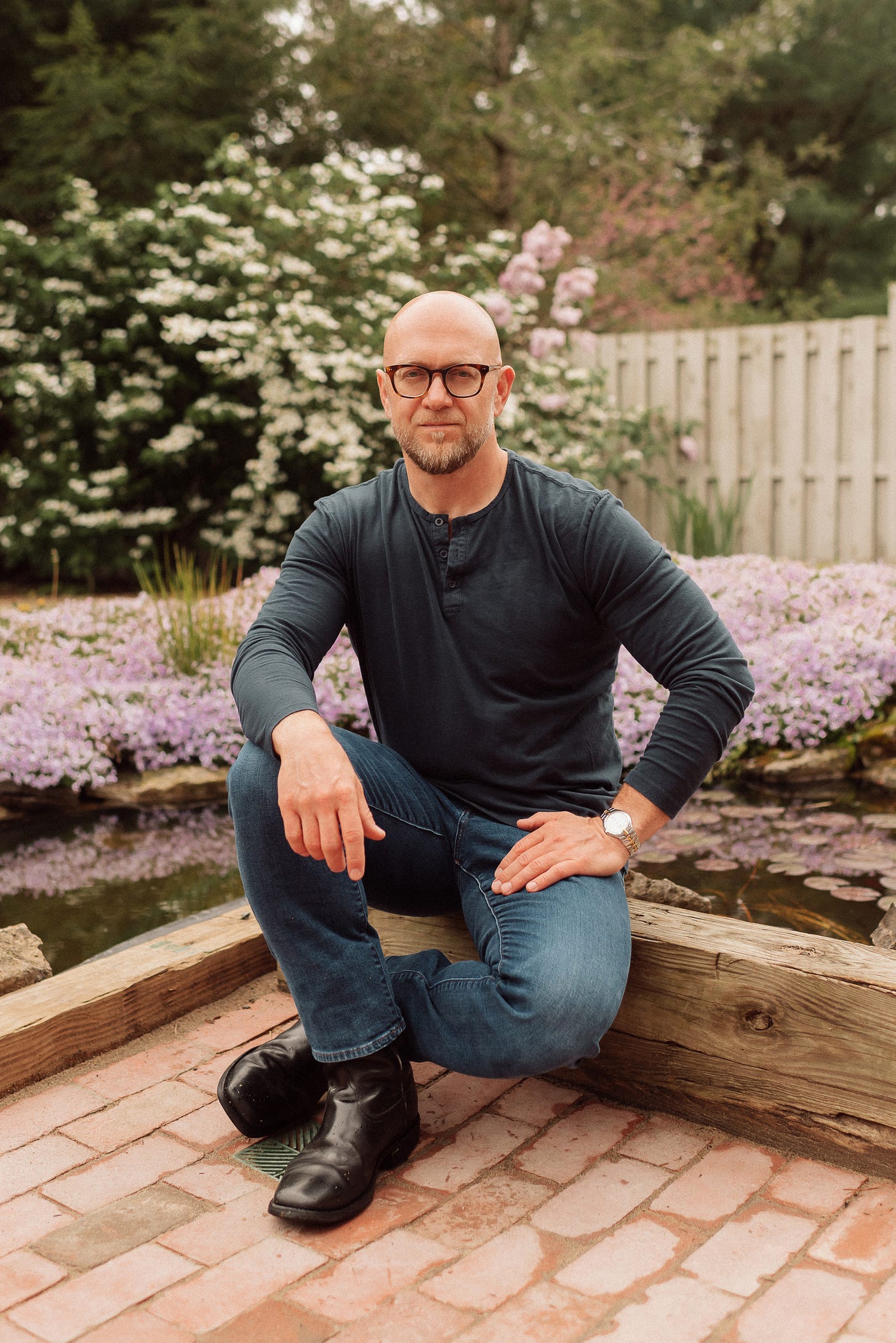 A bald man with a beard and glasses is kneeling on one knee in a garden. He is wearing a dark long-sleeved henley shirt, blue jeans, and black boots. He has a watch on his left wrist and is smiling slightly at the camera. Behind him, there are blooming pink and white flowers, a wooden fence, and lush green foliage, creating a serene and picturesque background. A bald man with a beard and glasses is kneeling on one knee in a garden. He is wearing a dark long-sleeved henley shirt, blue jeans, and black boots. He has a watch on his left wrist and is smiling slightly at the camera. Behind him, there are blooming pink and white flowers, a wooden fence, and lush green foliage, creating a serene and picturesque background.