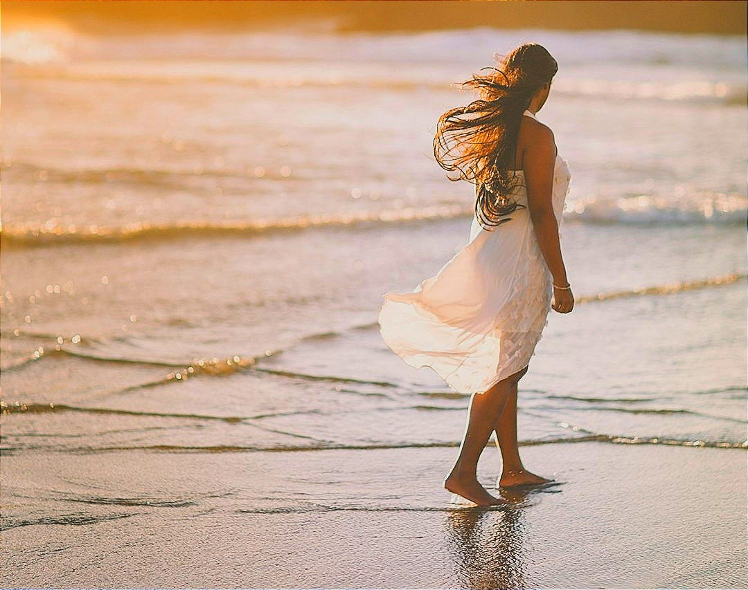 selective focus photo of woman standing on sea shore near rock formation during golden hour