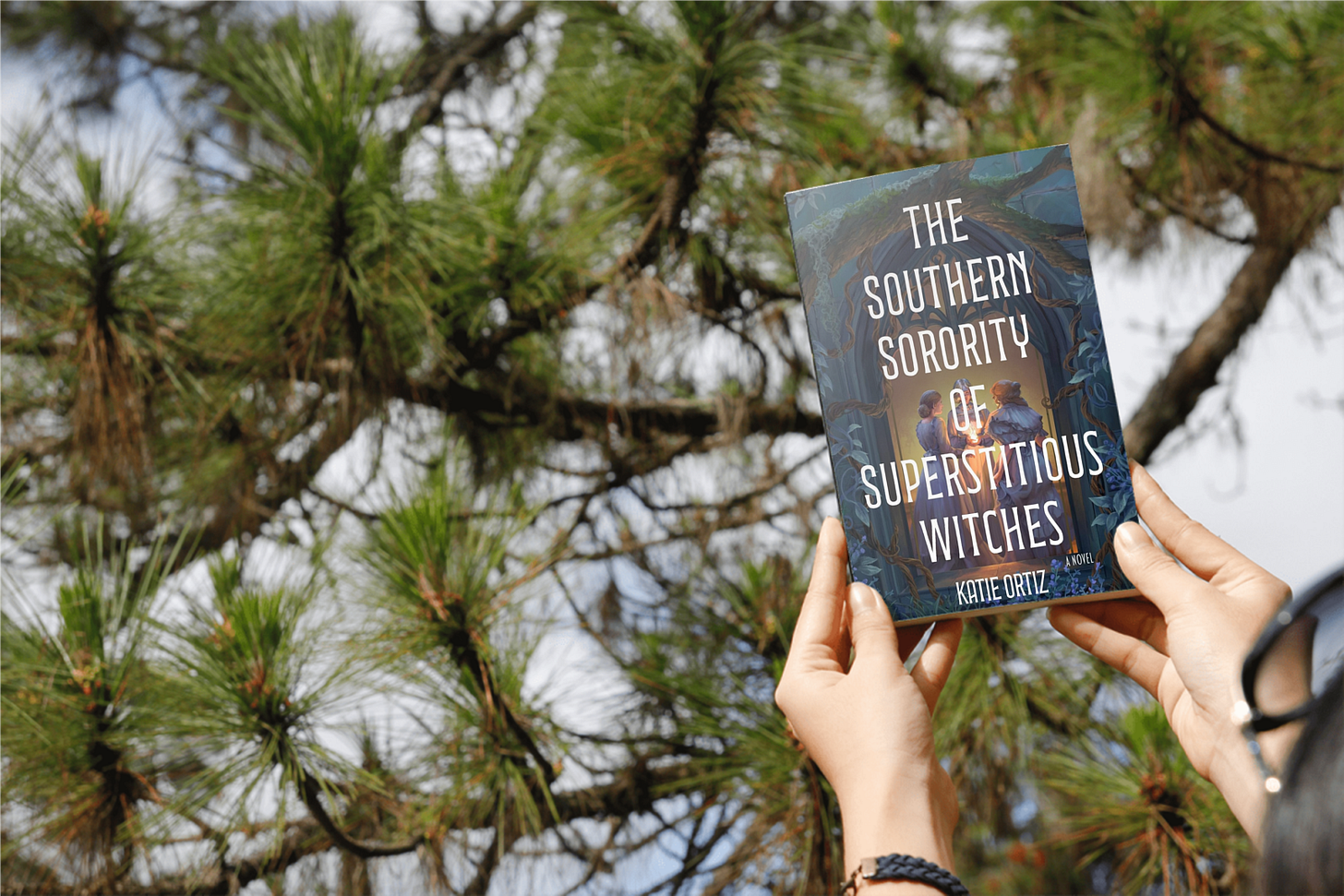 Book "The Southern Sorority of Superstitious Witches" by Katie Ortiz held by a women in front of a fir tree.