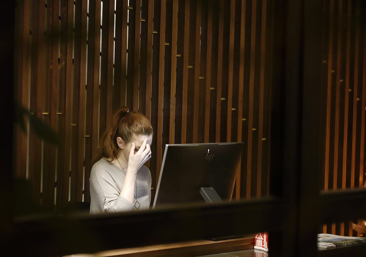 A woman sitting at a desk with computer in front of her. She is touching her forehead as though she is thinking or frustrated. In the background is a wall of brown wooden slats. A woman sitting at a desk with computer in front of her. She is touching her forehead as though she is thinking or frustrated. In the background is a wall of brown wooden slats.