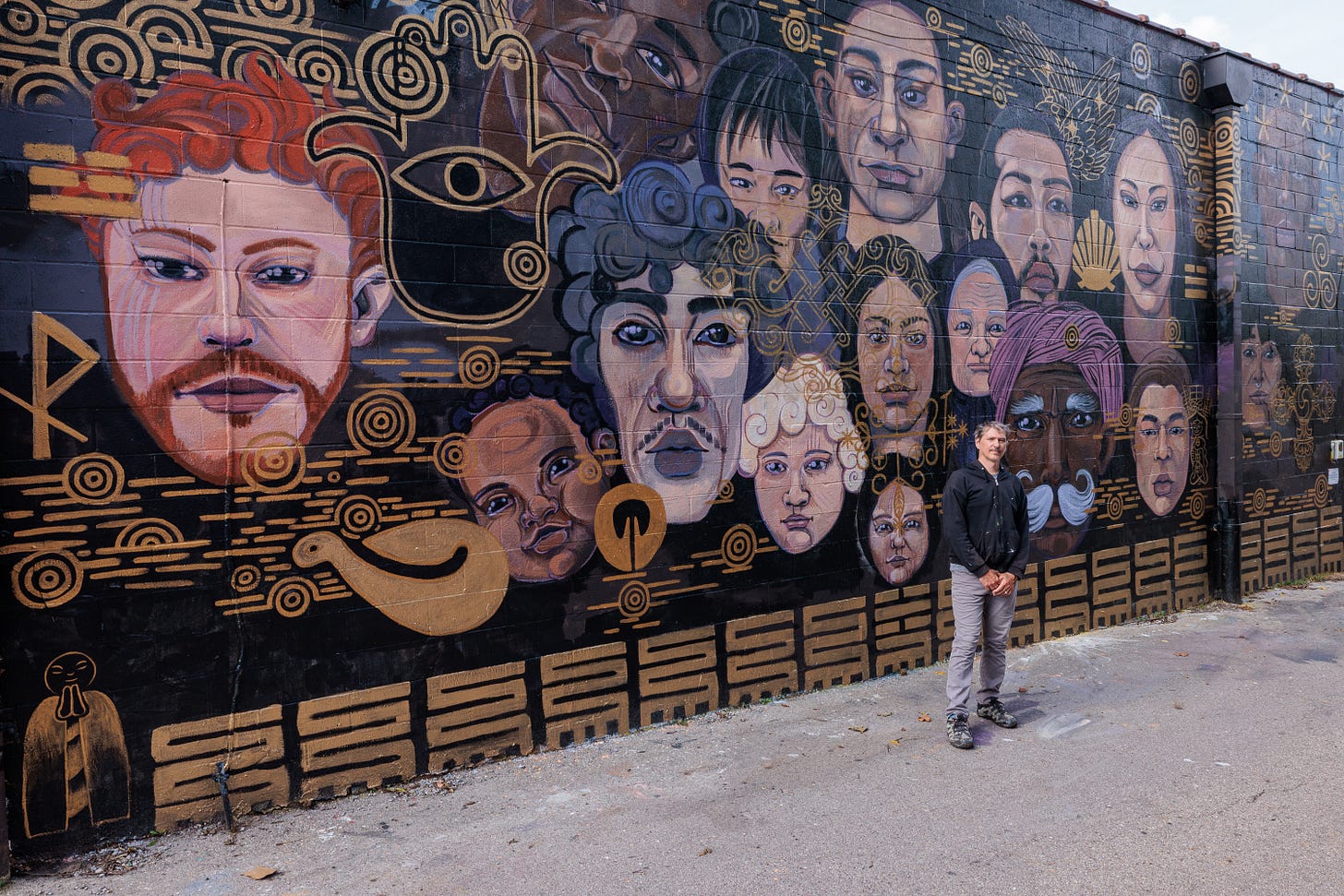 Pierre Nagley stands in front of the completed mural, a wall of portrait faces surrounded by gold linework and symbols on black.