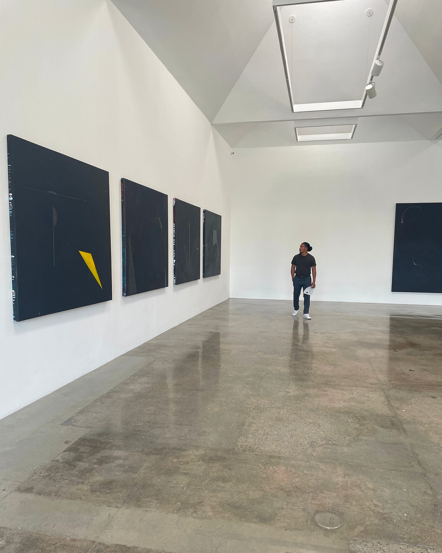 Man in an art gallery with while walls and polished concrete floors. He is wearing a dark green shirt, gray slacks, and white sneakers. He is walking toward the camera while looking over his right shoulder at four large oil paintings mounted on the wall. Man in an art gallery with while walls and polished concrete floors. He is wearing a dark green shirt, gray slacks, and white sneakers. He is walking toward the camera while looking over his right shoulder at four large oil paintings mounted on the wall.