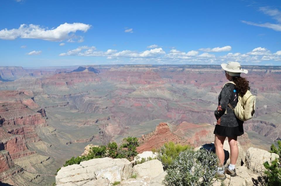 Claire olhando para a vastidão do Grand Canyon, sob um céu claro, transmitindo uma sensação de imensidão e silêncio.