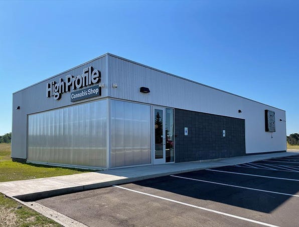 A modern cannabis shop building with a large sign, featuring a glass front and a parking lot under a clear blue sky.