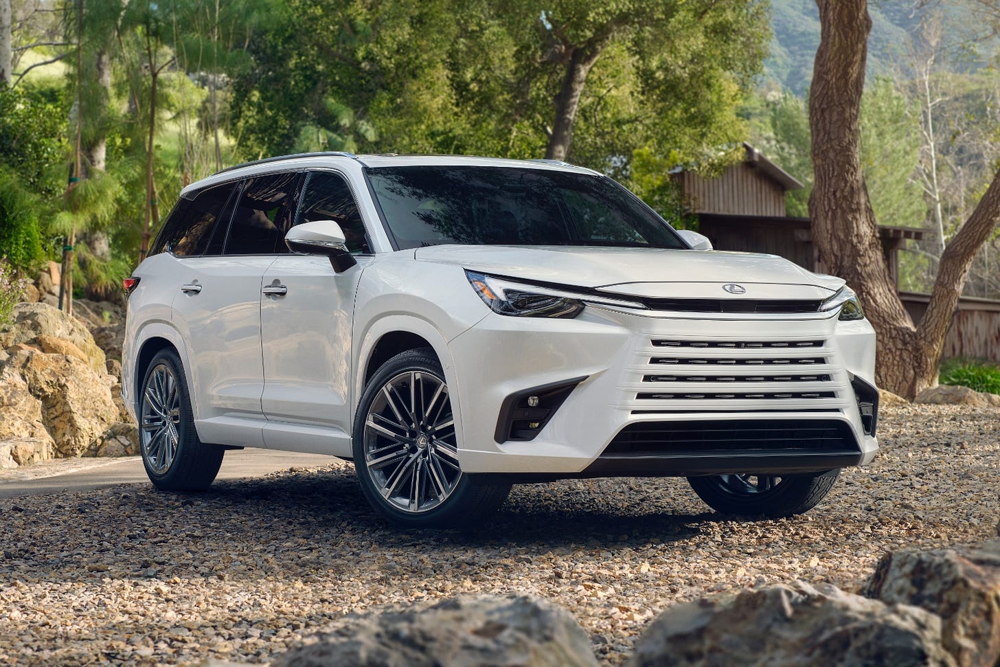 A white 2024 Lexus TX is parked on gravel outside of a cabin in the woods.