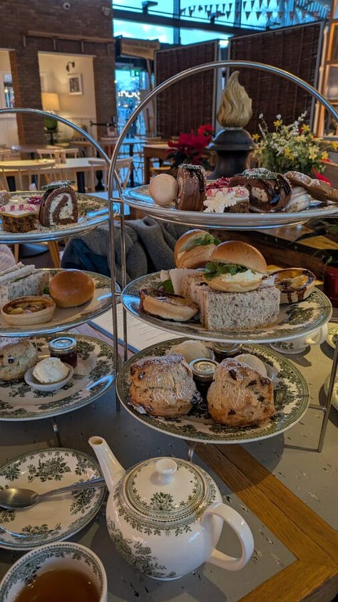 The first image shows a traditional British afternoon tea setup with tiered trays of scones, sandwiches, pastries, and desserts, along with a teapot and teacup on a floral-patterned table setting in a cozy café. The second image depicts a stone pathway leading toward the entrance of a grand historic castle tower surrounded by greenery. The third image shows another view of the same castle, focusing on a large round tower with a flag on top, stone walls, and manicured lawns bordered by a rope barrier.