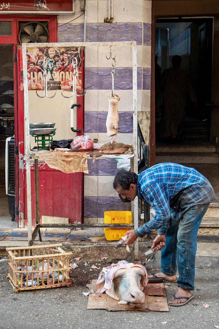 Top left: A narrow street in Luxor with apartment buildings, parked cars, and hanging textiles, with a Coptic church dome visible above the rooftops Top right: A woman in a black abaya buying fruit from a donkey-drawn cart loaded with oranges and bananas, parked in front of shuttered shopfronts Bottom right: A young man sitting inside a small vegetable stall framed by a green wall covered in Arabic calligraphy and religious posters, with piles of tomatoes, eggplant, and potatoes in the foreground Bottom left: A butcher in a blue plaid shirt bending over a cow's head on a cutting board, with a leg hanging from a hook and a Champions football poster on the red door behind him
