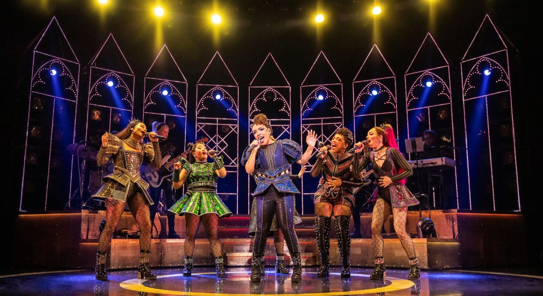 A group of women stood on stage wearing jewelled dresses and fishnets singing, with LED window arches behind them