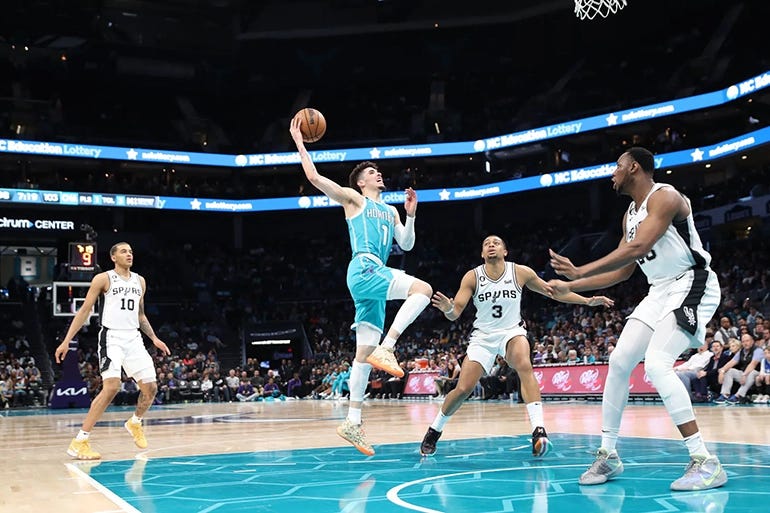Wearing a teal Charlotte Hornets jersey, LaMelo Ball extends himself while leaping toward the basket, holding the ball above his head and surrounded by three Spurs players. 