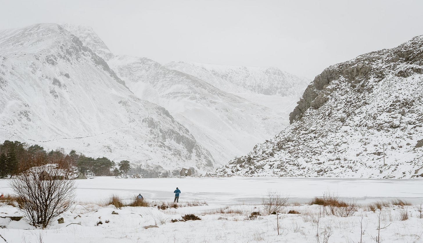 A person stands at the edge of a frozen lake. There are mountains in the distance.