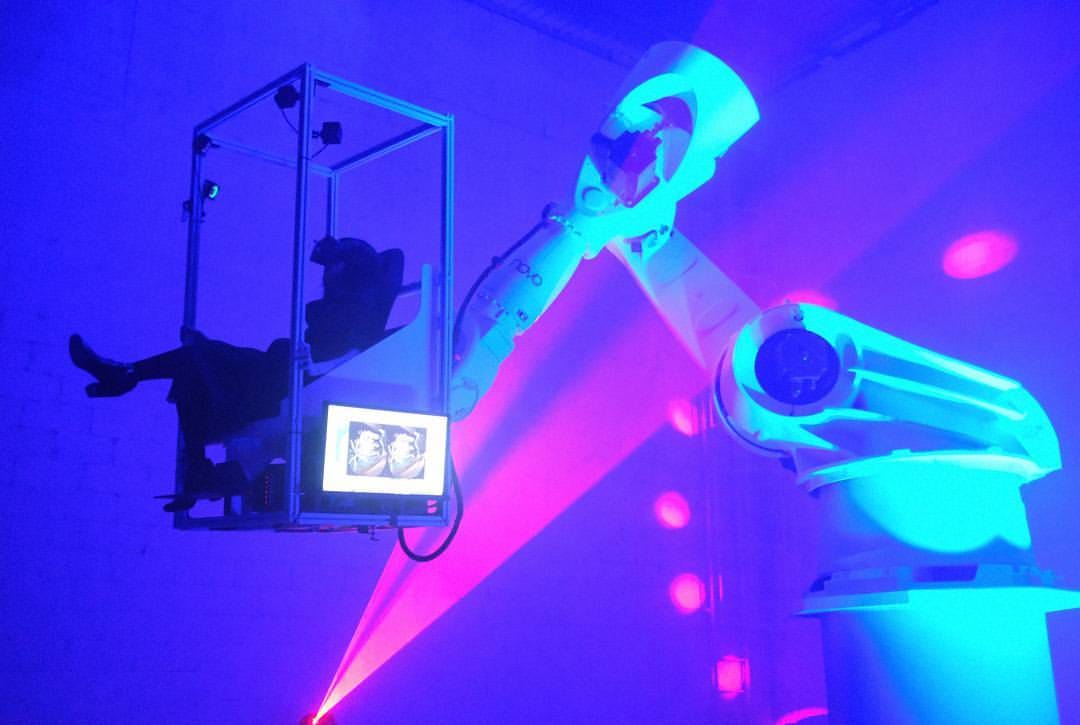 A large robotic arm, illuminated in vibrant blue and pink lighting, lifts a person seated in a suspended metal frame, creating a futuristic and immersive scene
