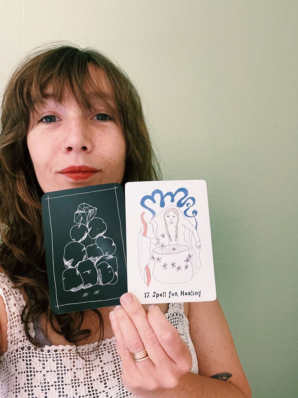 Casey Zabala holds up two cards, one from the Wanderer's Tarot deck is black and features an illustration of 10 stones, the other is a white card from her Wyrd Sisters Oracle deck and features an illustration of a figure standing over a cauldron, with a blue plume of smoke surrounding the illustration. Casey Zabala holds up two cards, one from the Wanderer's Tarot deck is black and features an illustration of 10 stones, the other is a white card from her Wyrd Sisters Oracle deck and features an illustration of a figure standing over a cauldron, with a blue plume of smoke surrounding the illustration.