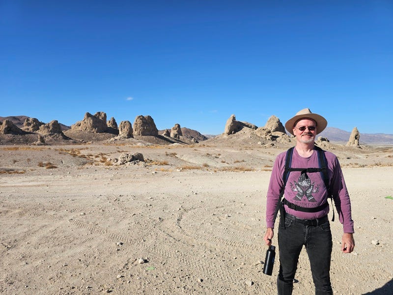 Man wearing a wide‑brimmed hat, sunglasses, backpack straps visible, and holding a water bottle, standing in a barren desert with cone‑shaped rock pinnacles in the distance under a clear blue sky.