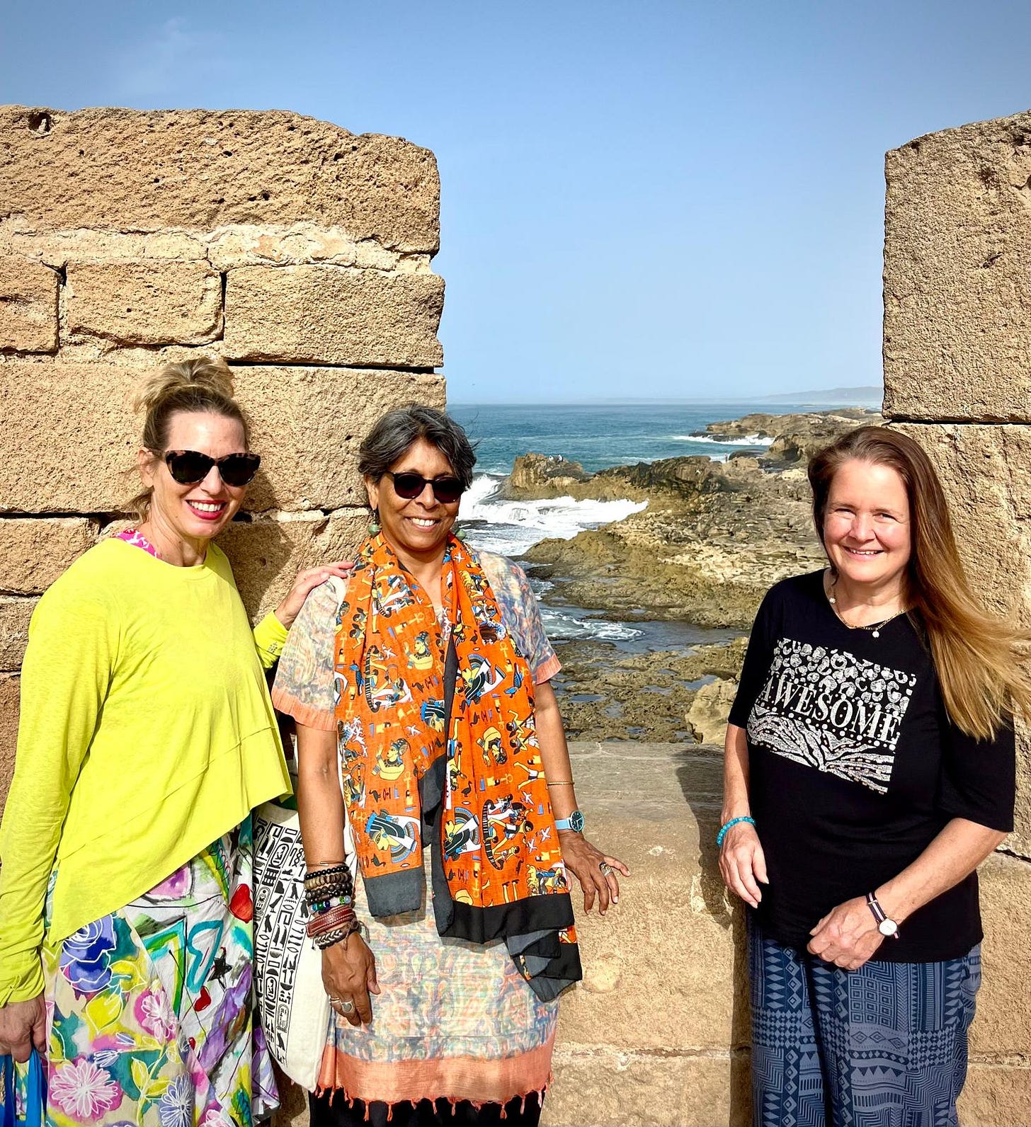 Two white women and a South Asian woman are wearing brightly colored clothes and standing in front of a rampant overlooking the ocean.    