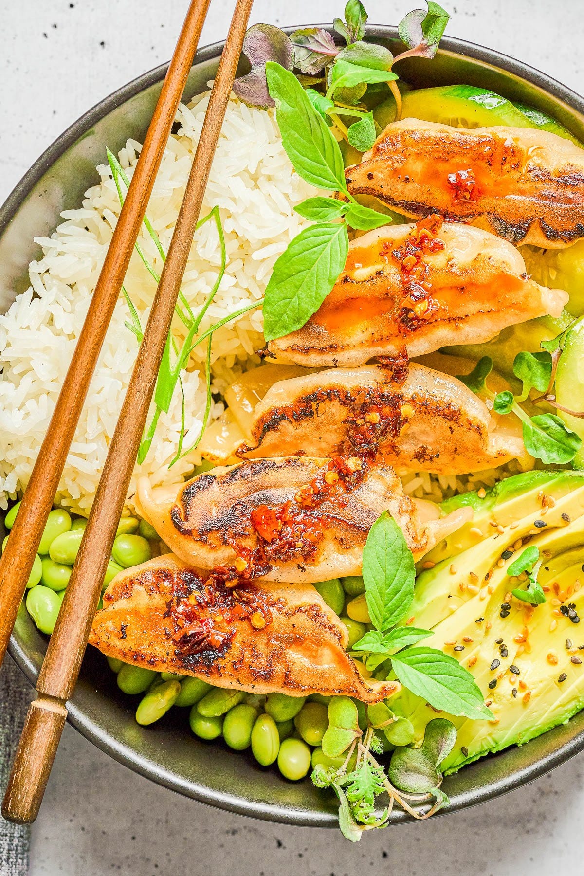 A bowl with steamed rice, five pan-fried dumplings topped with chili sauce, edamame, avocado slices, and fresh herbs, with wooden chopsticks resting on the bowl.