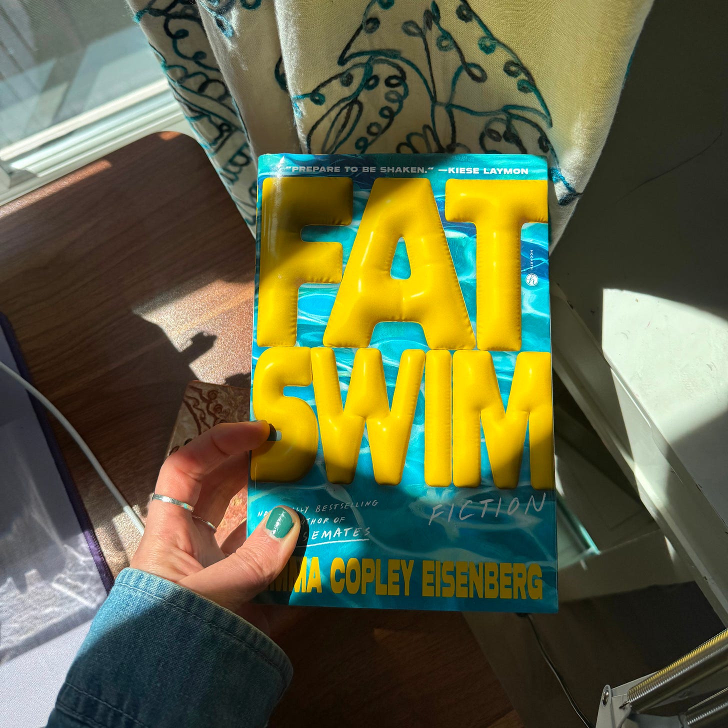 My hand holding my copy of Fat Swim over my desk. The vivid blue book cover glistens in the sunlight coming in from the window behind it.