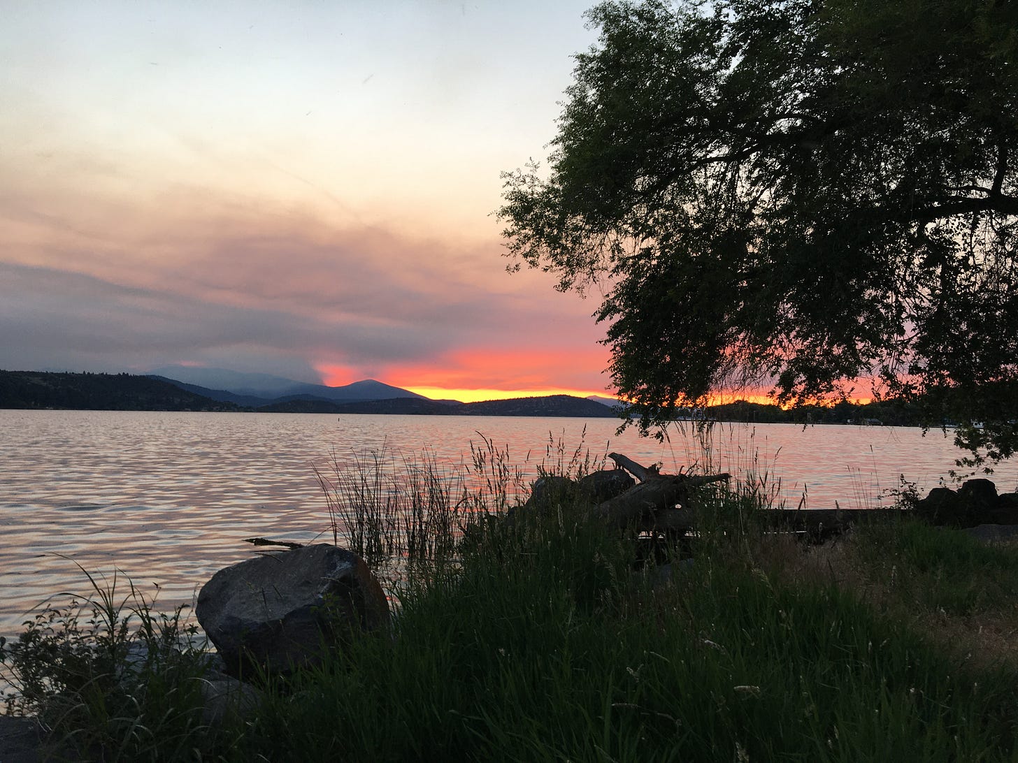 Sunset by a lake. Plumes of smoke mix with brilliant reds and yellows over the partially shrouded mountains. 
