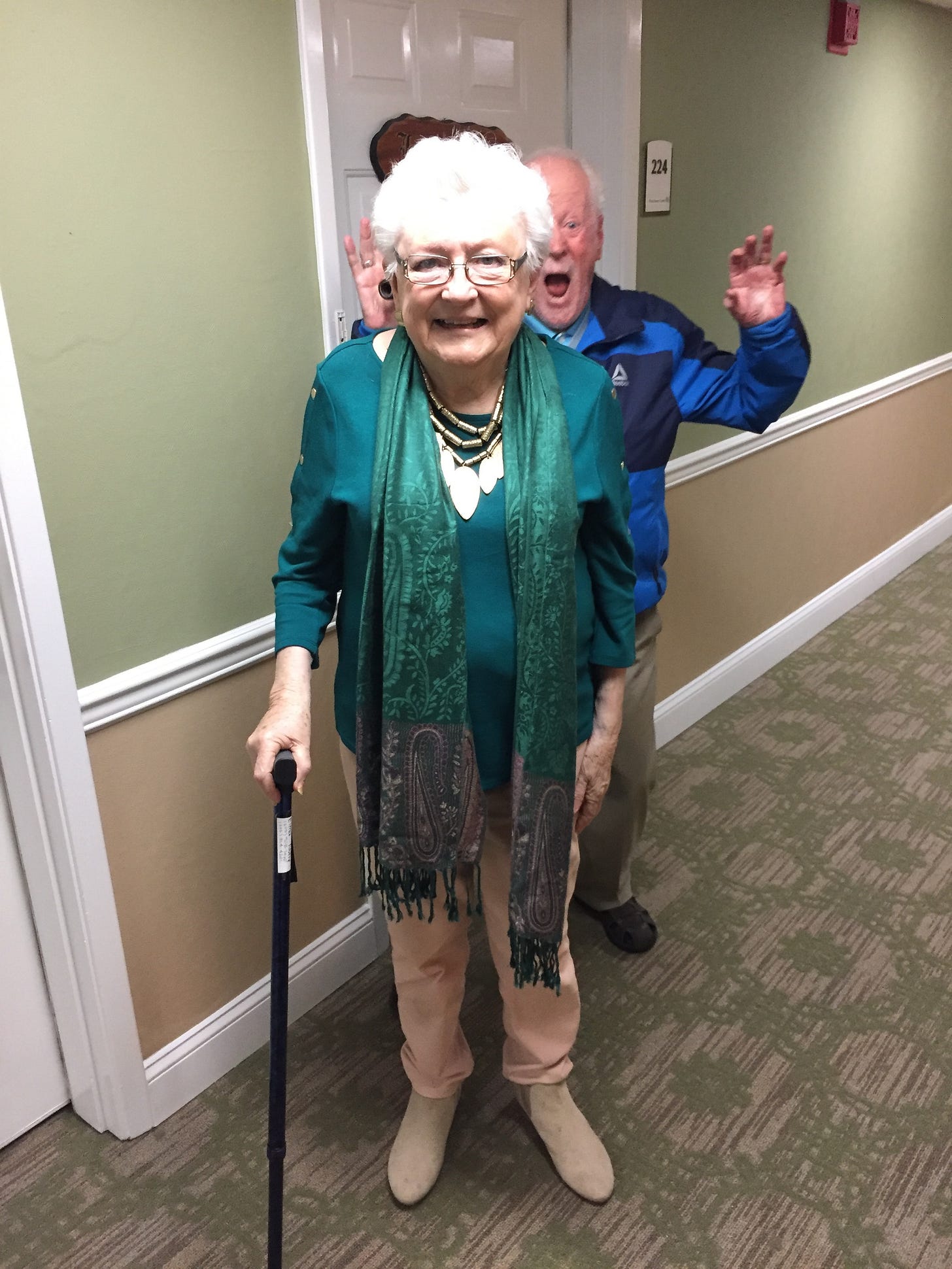 Older lady standing in a hallway with he husband making funny face and gestures behind her
